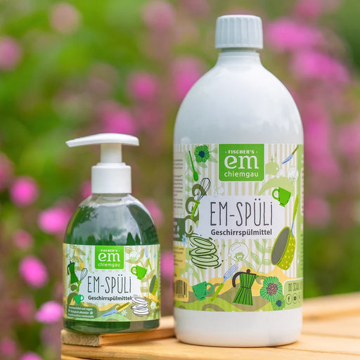 EM Chiemgau EM-Spüli Geschirrspülmittel als 250 ml Spenderflasche und 1 Liter Nachfüllflasche nebeneinander auf einer Holzoberfläche mit grünem und rosa Blumenhintergrund.
