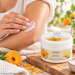 Bergland Melkfett mit Bio-Ringelblume 200 ml in weißer Cremedose auf einem Holzbrett, während eine Person die Creme auf einen trockenen, beanspruchten Ellenbogen aufträgt, umgeben von orangefarbenen Ringelblumen und weichen Handtüchern in natürlichem Pflegeambiente.