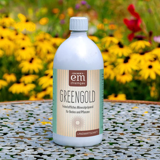 EM Chiemgau GREENGOLD 1 Liter, feinstoffliches Mineralpräparat für Boden und Pflanzen, in weißer Flasche mit hellgrünem Etikett auf einem Gartentisch, umgeben von leuchtend gelben Blüten und bunten Gartenpflanzen.