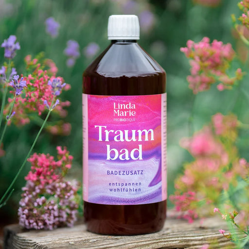 EM Chiemgau Traumbad 1 Liter Badezusatz in brauner Flasche mit rosa-violettem Etikett auf rustikalem Holztisch, umgeben von zarten rosa und violetten Blüten in einem natürlichen Gartenambiente.