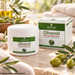 Golden Heart Olivenöl Körperbalsam 250 ml mit Vitamin E und Ringelblumenextrakt, weiße Cremedose Originalverpackung auf heller Steinoberfläche, umgeben von grünen Oliven, Olivenzweigen und weichen Handtüchern.