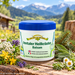 Inntaler Heilkräuter Balsam im weißen Tiegel mit blauem Deckel, umgeben von verschiedenen Kräutern und Blüten vor sonniger alpiner Berglandschaft.