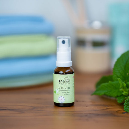 EMsana Zahnfit Spray 20 ml in brauner Glasflasche mit weißem Sprühkopf, auf einer Holzoberfläche mit frischen grünen Blättern im Badezimmerambiente.